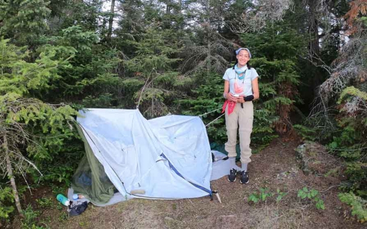 a person stands beside their tarp shelter in a wooded area on an outward bound trip
